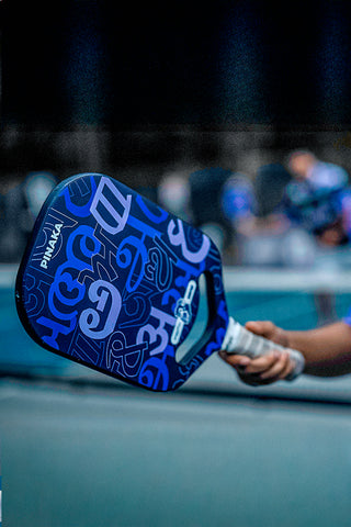 A close-up action shot of a pickleball player's hand holding a blue and purple patterned Pinaka paddle on a court.