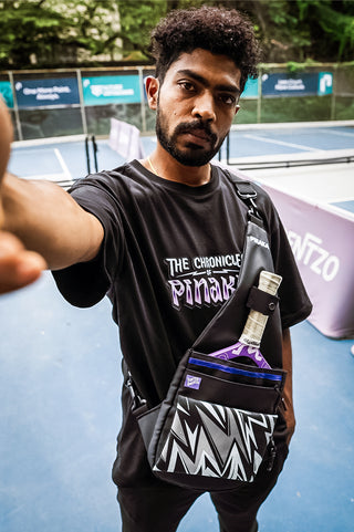 A man on a blue pickleball court wearing a black t-shirt and a black crossbody sling bag with a geometric patterned bottom pocket. The bag holds a pickleball paddle.