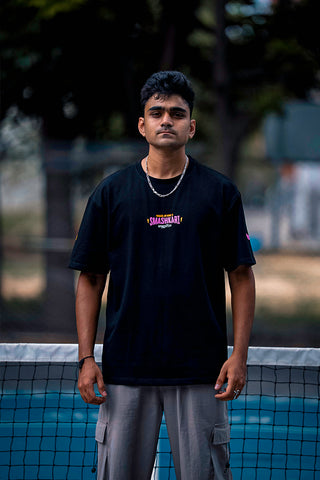 A man standing on a pickleball court wearing a black t-shirt with a colorful 'SMASHKARI' graphic.