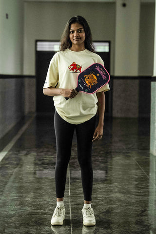 A woman standing in a hallway wearing a beige t-shirt holding a blue and pink pickleball paddle.